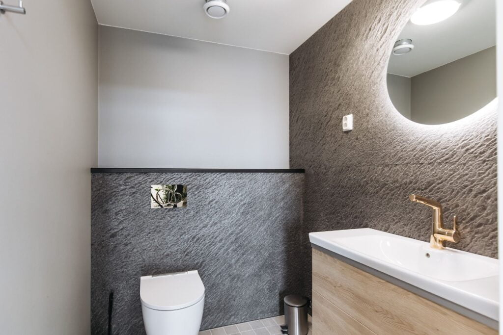 Pieni wc-tila, jossa pyöreä taustavalaistu valopeili ruskeasävyisellä seinällä allaskalusteen päällä