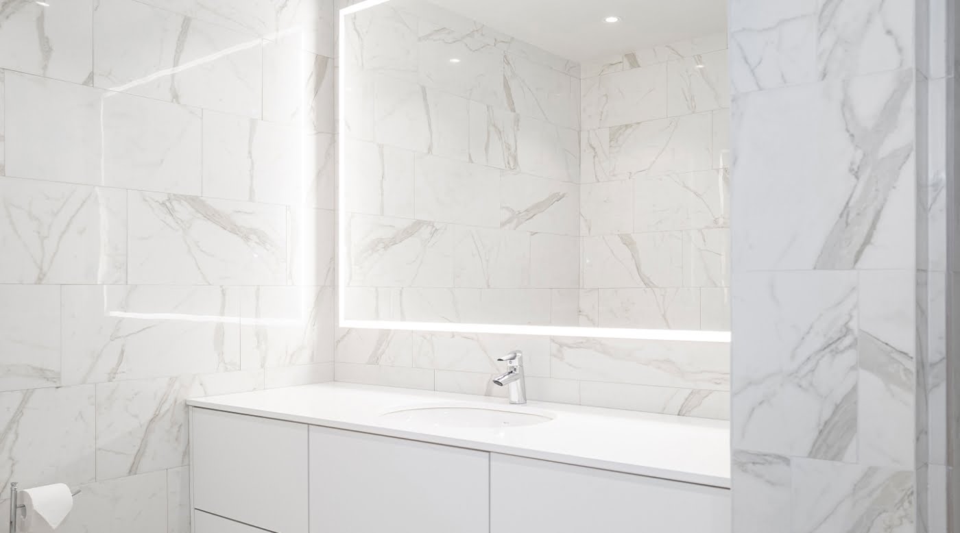 Vaaleasävyinen kylpyhuone marmorikuvioiduilla laatoilla. Keskellä iso valopeili pesualtaan yllä. Valopeili valaisee kauniisti koko kylpyhuoneen.